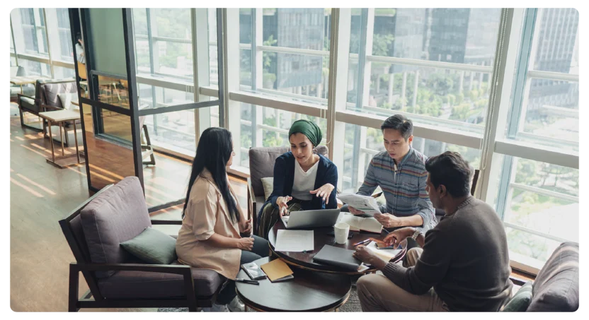 Four people sit around a coffee table in a modern office lounge with large windows, collaborating on work with a laptop, papers, and notebooks. Natural light fills the room, creating a bright atmosphere.