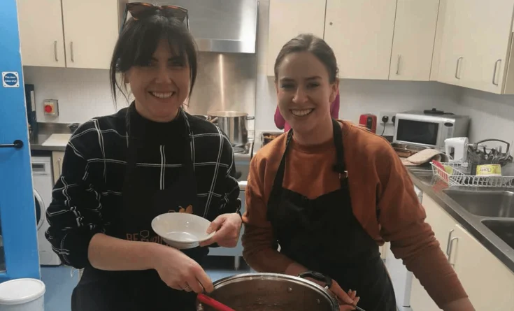 Two women standing in a kitchen, both wearing dark aprons and smiling at the camera. One holds a bowl and the other is stirring a large pot of food on the counter. Kitchen appliances are visible in the background.