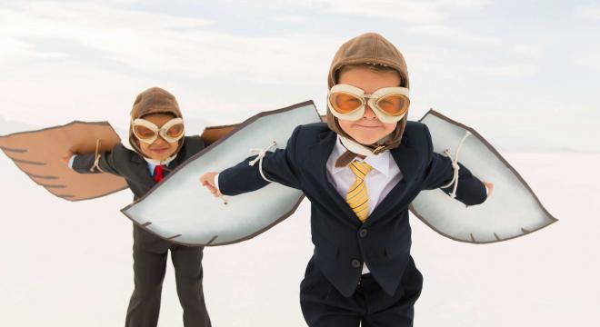 Two children wearing suits, aviator hats, goggles, and large cardboard wings pretend to fly while standing on a flat, open white surface under a bright sky.