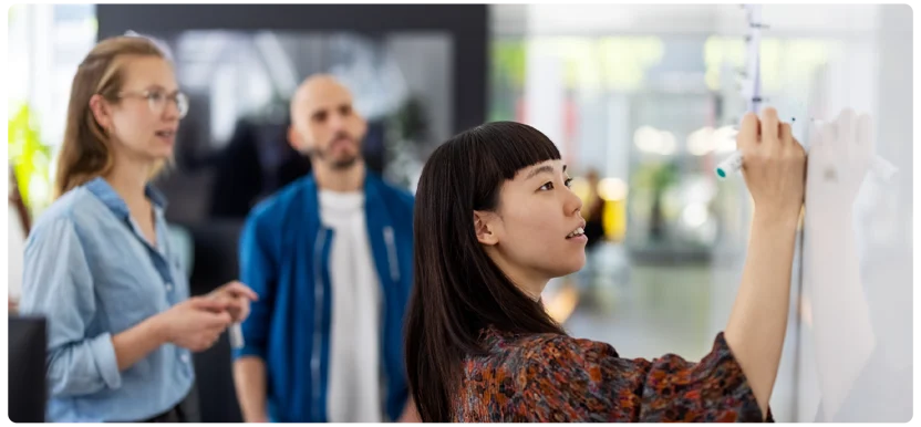 A woman writes on a whiteboard with a marker, while two colleagues stand nearby watching her in a bright, modern office setting.