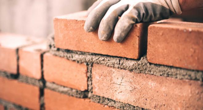 A gloved hand carefully places a red brick onto a wall under construction, with fresh mortar visible between the bricks.