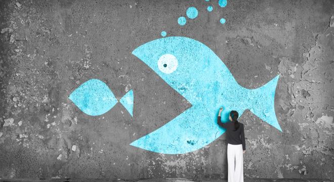 A woman draws on a wall featuring graffiti of a large blue fish about to eat a smaller blue fish, with blue bubbles above the big fish’s head.