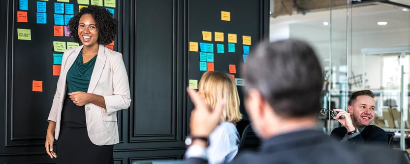 A woman stands smiling and presenting in front of a wall covered with colorful sticky notes, while colleagues seated at a table listen and engage with her in a modern office setting.