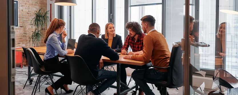 Six people sit around a conference table in a modern office, engaged in discussion. Glass walls, large windows, and indoor plants create a bright, collaborative atmosphere.