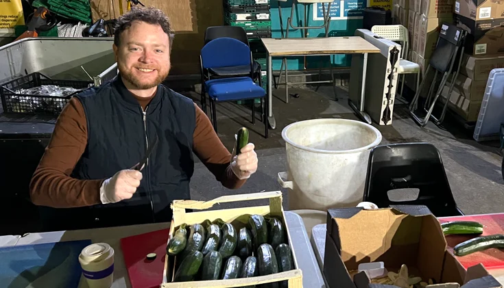 A smiling man wearing gloves sits at a table, holding a knife and a cucumber. Several cucumbers are in a box in front of him, and various containers and supplies are on the table. The setting appears to be a kitchen or prep area.