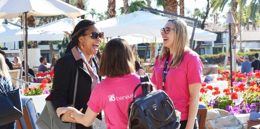 Three people laugh and chat together outdoors at a sunny event, two wearing bright pink “Benevity” shirts, surrounded by flowers, palm trees, and white umbrellas.
