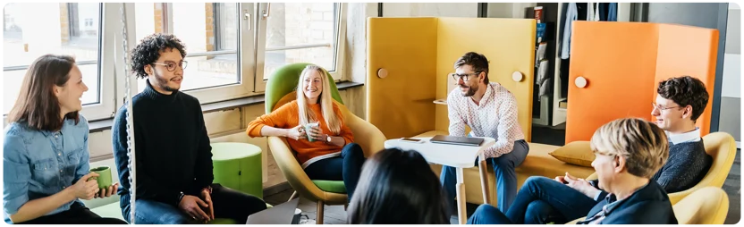 A group of seven people sit in a circle on colorful chairs in a modern, bright office space, smiling and talking with each other in a casual meeting.