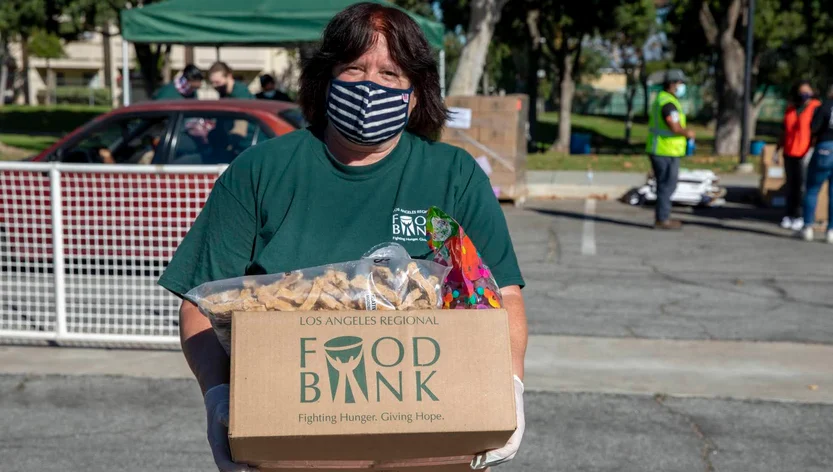 A woman wearing a striped mask and green shirt holds a box labeled Los Angeles Regional Food Bank filled with food items at an outdoor distribution event.