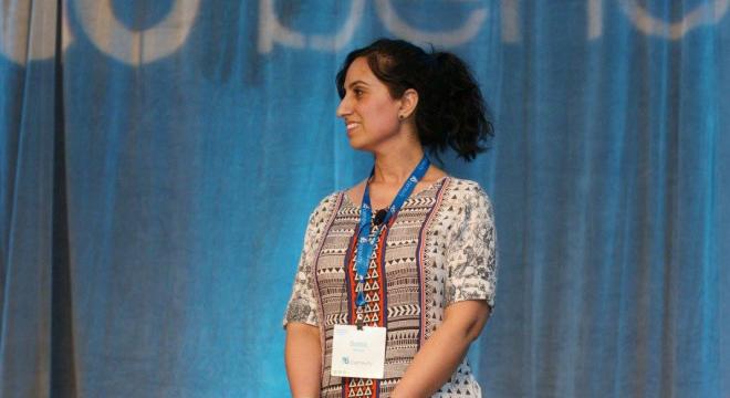 A woman with dark hair in a ponytail stands on stage, smiling to the side. She is wearing a patterned dress and a blue lanyard with a badge, with a blue curtain and blurred text in the background.