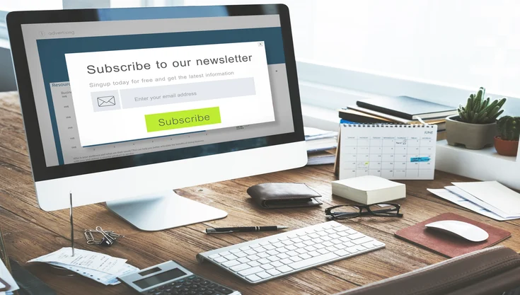 A computer monitor on a desk displays a “Subscribe to our newsletter” pop-up. The workspace includes a keyboard, mouse, notepad, pen, calendar, documents, and a mug.
