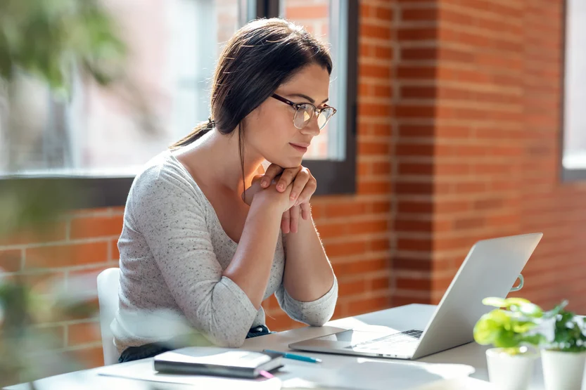 A woman with glasses sits at a desk, looking thoughtfully at her laptop. Sunlight streams through large windows, and notebooks, a pen, and a small plant are on the desk beside her.