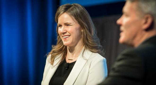 A woman in a light-colored blazer is smiling while talking to a man in a dark suit. The background is blurred with blue and gray tones, suggesting a professional or conference setting.