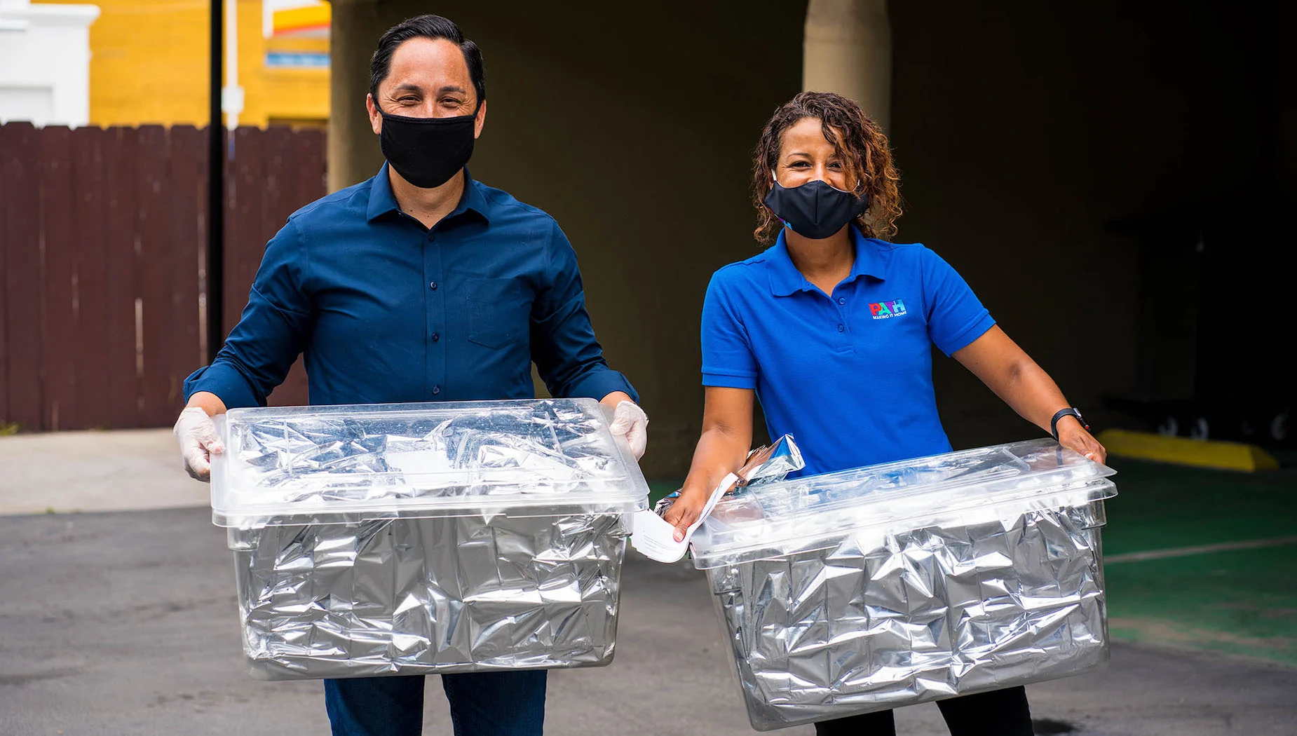 Two people wearing face masks and gloves stand outdoors, smiling and holding large clear plastic bins wrapped in silver insulation. They appear to be participating in a distribution or delivery event.
