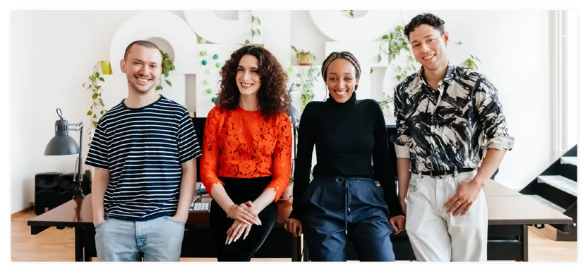 Four people stand side by side in a modern office, smiling. There are plants in the background and desks behind them. They are casually dressed and appear friendly and relaxed.