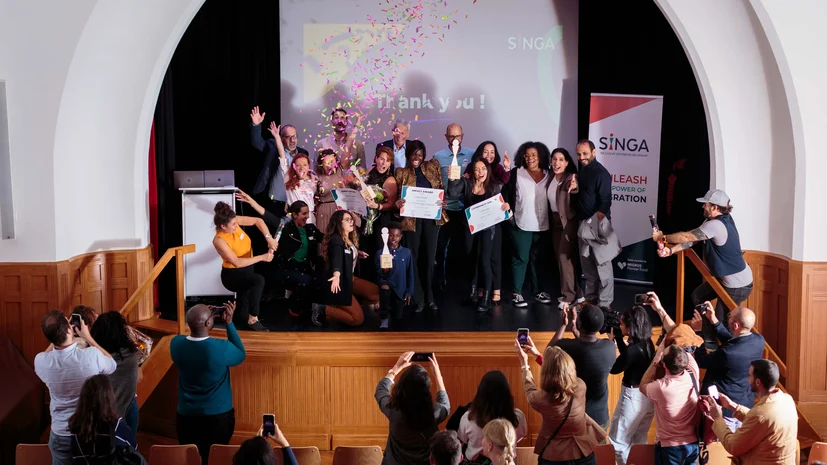 A group of people celebrate on stage holding certificates as confetti falls, while an audience takes photos. A screen behind them displays “Thank you!” and banners show the SINGA logo.