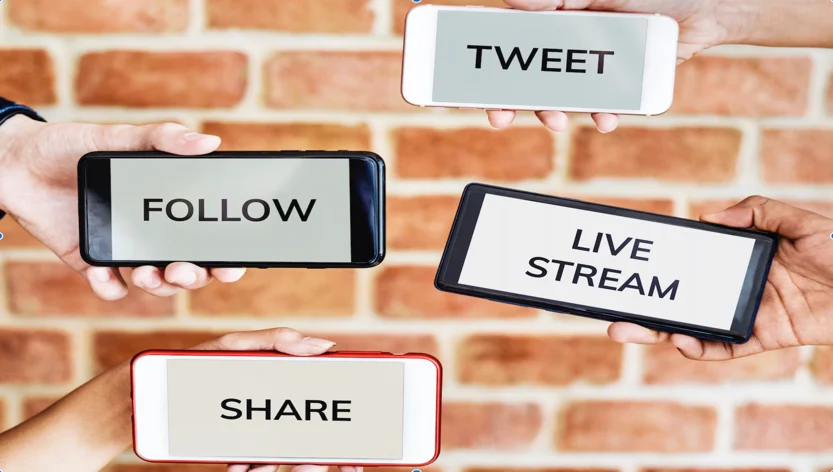 Four hands holding smartphones displaying words: TWEET, FOLLOW, LIVE STREAM, and SHARE, against a brick wall background.