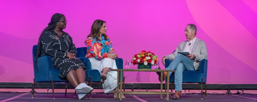 Three people sit on stage in armchairs, engaged in a panel discussion. Two women sit on the left, and a man sits on the right holding a tablet. A table with a flower arrangement is between them, against a pink backdrop.
