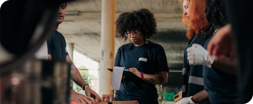 A group of volunteers wearing dark shirts work together at a table, one person holding a clipboard and paper while others sort and organize items, possibly at a community service event.