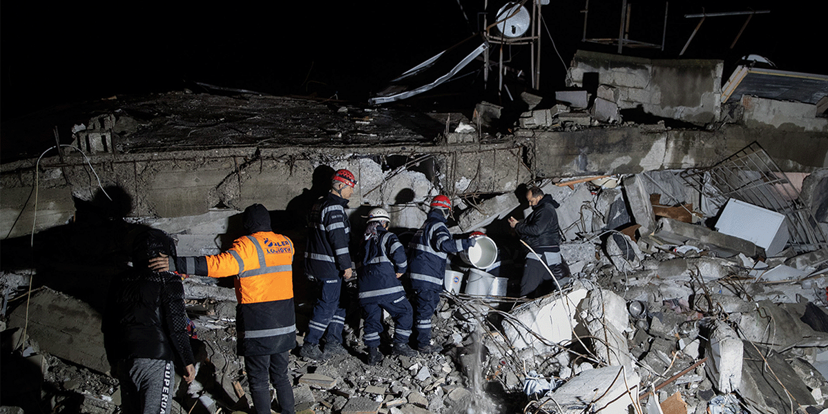 Rescue workers in safety gear search through the rubble of a collapsed building in Türkiye at night, using flashlights and tools, with debris and broken structures surrounding them.