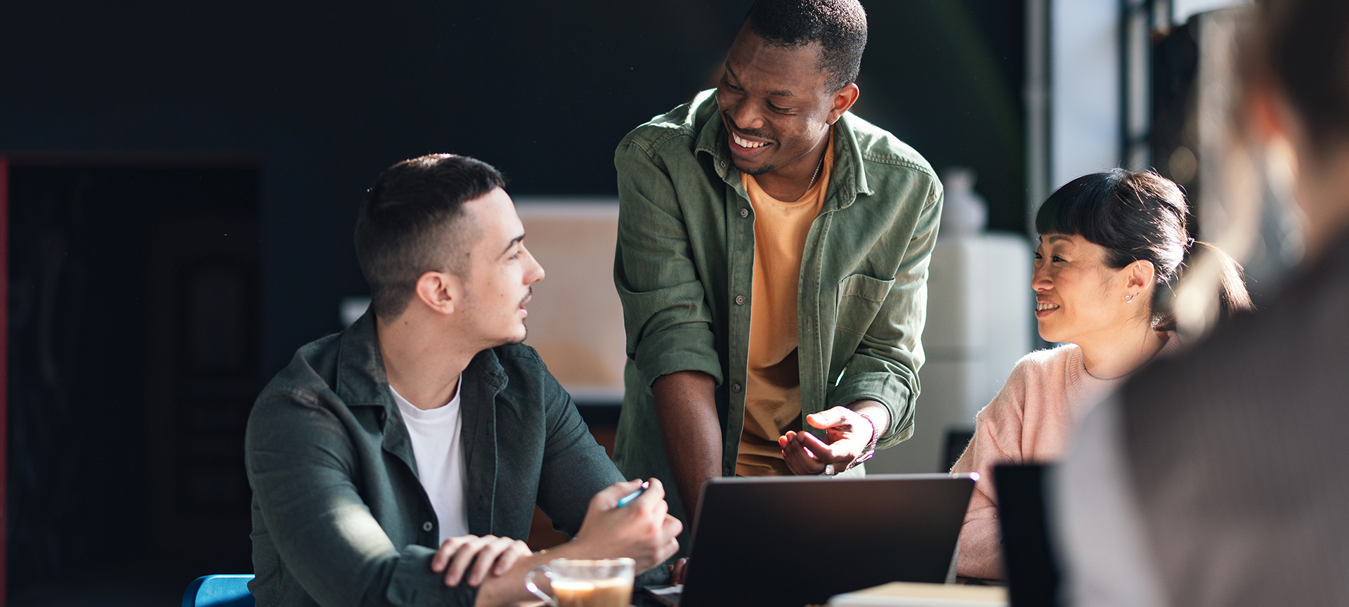 Three people sit at a table with a laptop and coffee, engaged in a friendly conversation. One person stands and smiles, while the other two sit and listen attentively in a bright, modern office setting.