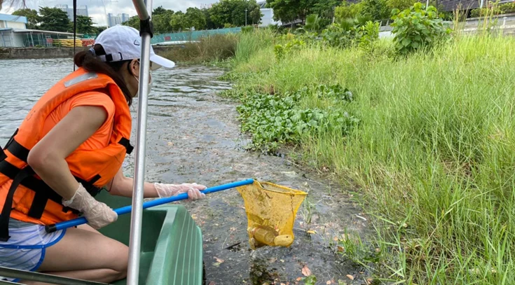 A person wearing an orange life vest and white cap cleans a lakeshore with a yellow net from a boat, collecting debris floating near grassy waters edge.