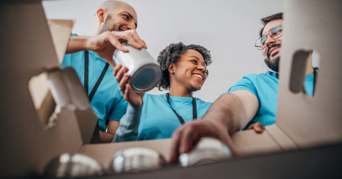 Three people in blue shirts smile and work together, placing canned goods into a large cardboard box, viewed from inside the box.
