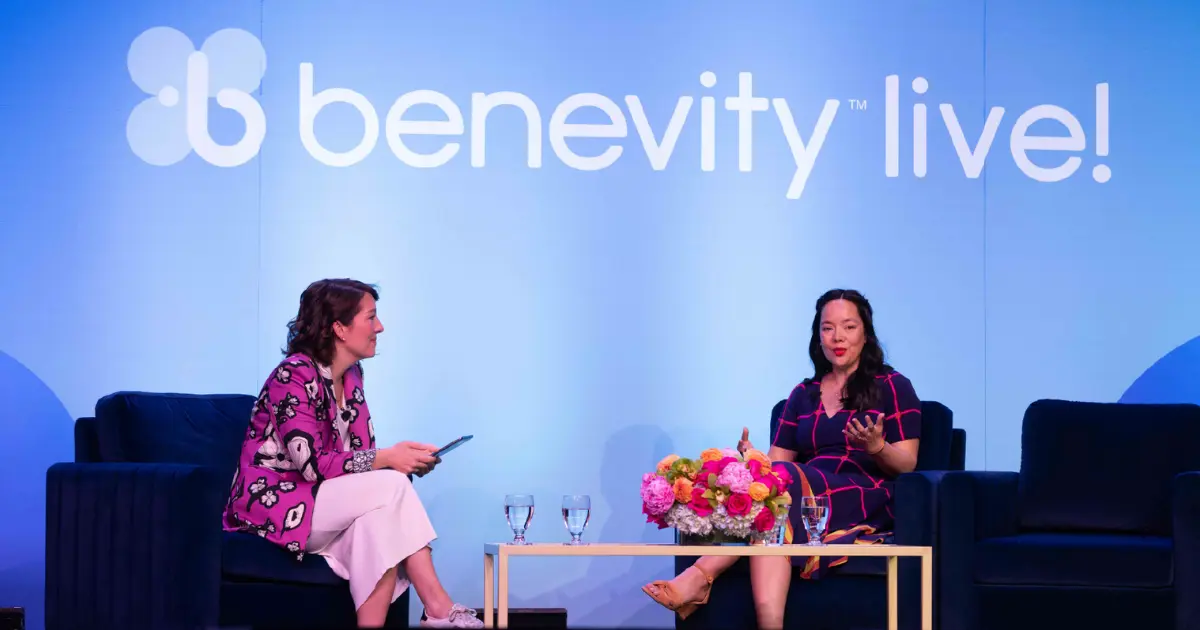 Two people sit on stage in front of a “Benevity Live!” sign, talking. One holds a tablet, the other gestures while speaking. They are seated on dark chairs with a table between them, holding flowers and water glasses.