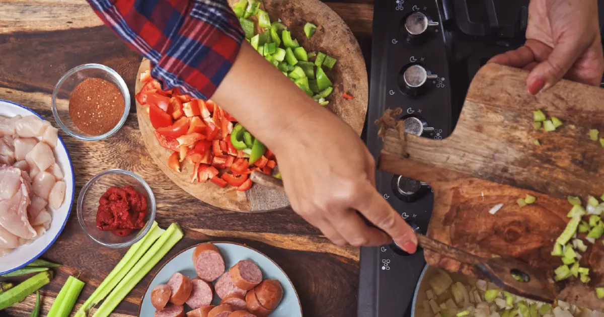 A person chops and adds vegetables to a pan on a stove, surrounded by bowls of chopped chicken, sausage, spices, celery, and peppers on a wooden countertop.