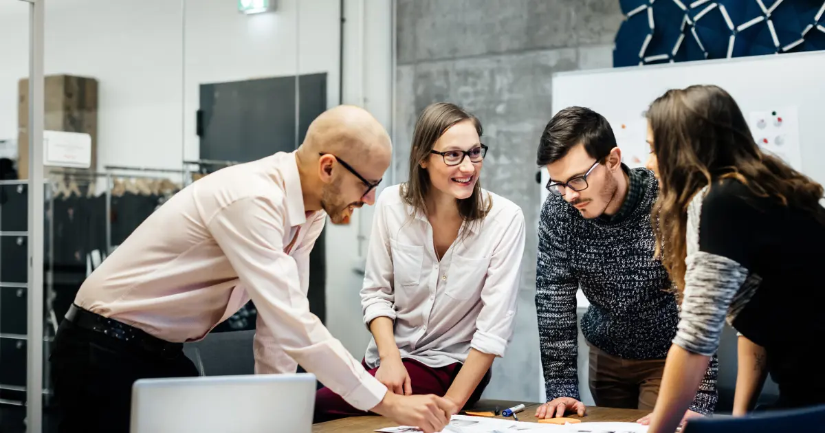 Four people gather around a table, engaged in discussion and looking at documents, suggesting a collaborative work or brainstorming session in a modern office setting.