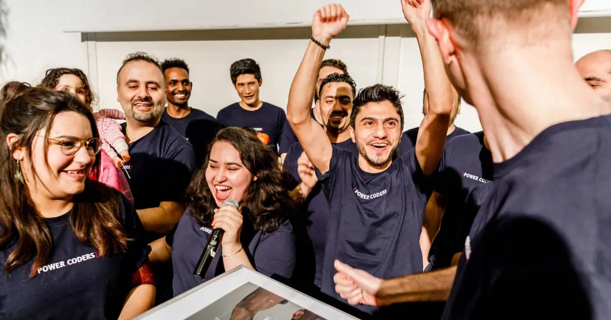 A group of people wearing matching Power Coders shirts celebrate together indoors. Some are smiling, one person holds a microphone, and another raises their arms in excitement.