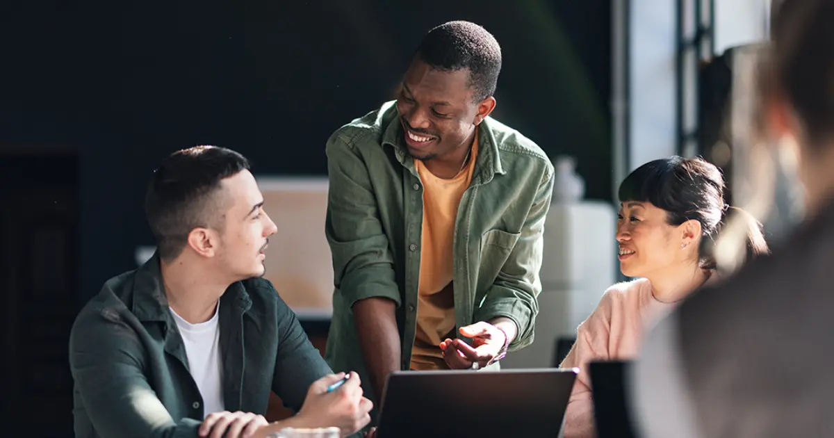 Three people sit at a table with a laptop and coffee, engaged in a friendly conversation. One person stands and smiles, while the other two sit and listen attentively in a bright, modern office setting.