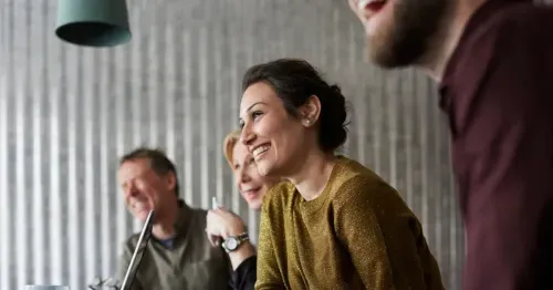 Four adults sit at a table in a bright room, smiling and laughing together. A laptop and coffee mug are visible on the table, and a green hanging lamp is above them. The mood is cheerful and collaborative.