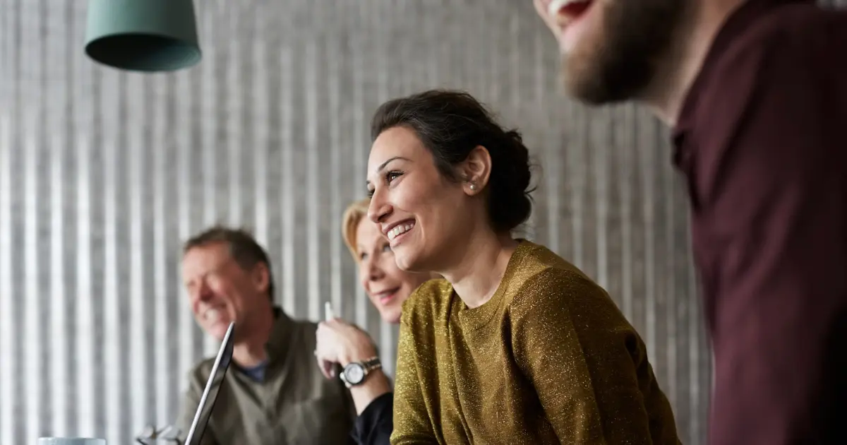 Four adults sit at a table in a bright room, smiling and laughing together. A laptop and coffee mug are visible on the table, and a green hanging lamp is above them. The mood is cheerful and collaborative.