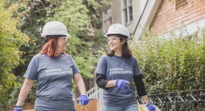 Two women wearing hard hats, gloves, and matching energizing COMMUNITIES shirts smile and walk together outdoors near a building, appearing to participate in a community service or volunteer activity.