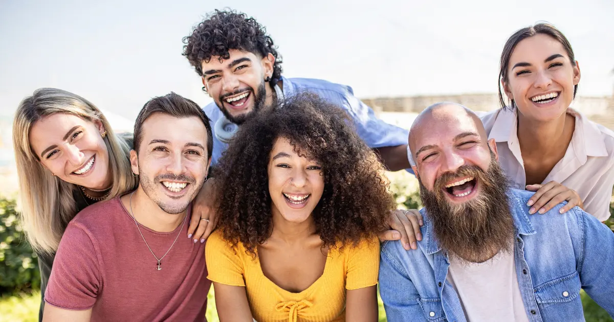 A group of six young adults, smiling and laughing together outdoors on a sunny day, sitting closely and appearing happy and relaxed.