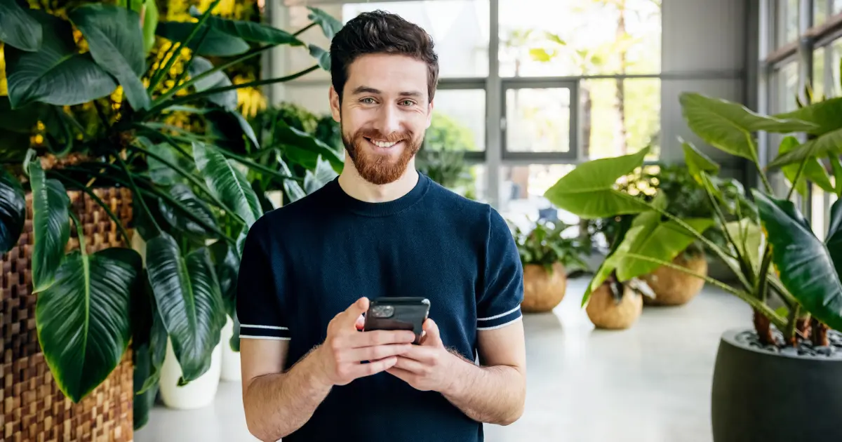 A smiling man with a beard stands indoors among large green plants, holding a smartphone and looking at the camera.