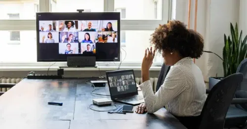 A person sits alone at a conference table, participating in a video call on a laptop, with a larger screen showing many people in a virtual meeting. A phone and other devices are on the table.