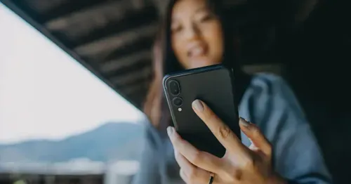 A woman holding a smartphone and smiling, with her face slightly out of focus. She is wearing a blue sweater and a watch, and is standing outdoors under a roof with mountains in the background.