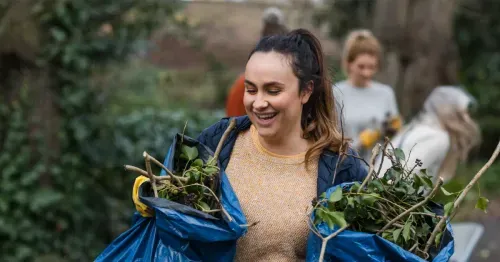 A smiling woman carries blue bags filled with green branches and leaves, while other people work in the background in an outdoor, wooded area.