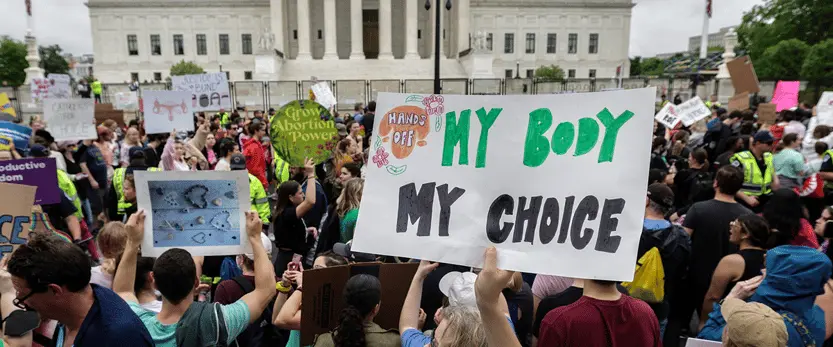 A large crowd protests outside a government building, holding various signs including one in front that reads “My Body My Choice.” Other signs on reproductive rights are visible among the crowd.