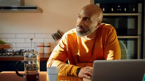 A man in an orange sweater sits at a kitchen table with a laptop, coffee cup, and French press, looking thoughtfully towards the window as sunlight streams in.