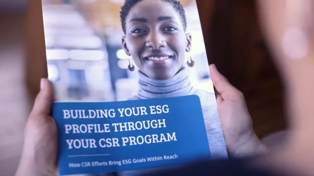 A person holds a brochure featuring a smiling woman on the cover and the text BUILDING YOUR ESG PROFILE THROUGH YOUR CSR PROGRAM with a subheading How CSR Efforts Bring ESG Goals Within Reach.