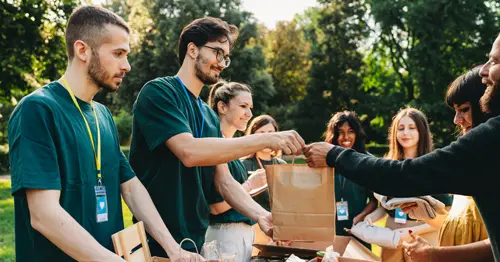 A diverse group of volunteers in green shirts distribute food and supplies outdoors, handing paper bags and packaged goods to people across a table, with trees visible in the background.