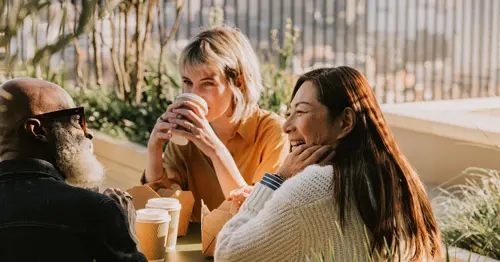 Three people sit at an outdoor table, smiling and drinking coffee together. Sunlight highlights their faces, and there are coffee cups and takeaway boxes on the table, with greenery in the background.