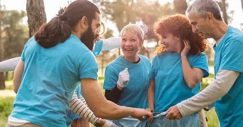 A group of smiling people wearing blue shirts gather outdoors, holding a large trash bag and picking up litter in a park, with trees and sunlight in the background.