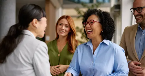 Four professionally dressed people greet each other indoors. Two women, one with curly hair and glasses, shake hands while smiling. Two others, a woman and a man, stand nearby, watching and smiling.