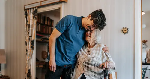A young man in a blue polo shirt hugs and smiles with an elderly woman seated in a chair, both appearing joyful in a cozy living room setting.