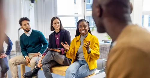 A group of young adults sits in a circle indoors, engaged in discussion. One woman in a yellow jacket speaks enthusiastically while others listen attentively, creating an atmosphere of collaboration and communication.