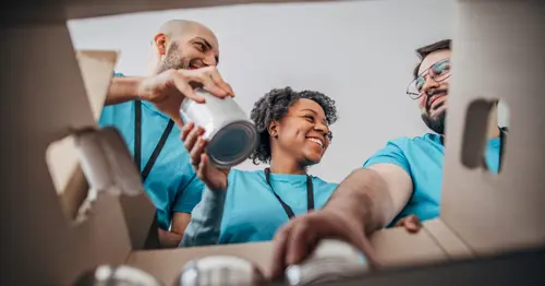 Three volunteers in blue shirts smiling and packing food cans into a cardboard box, viewed from inside the box.