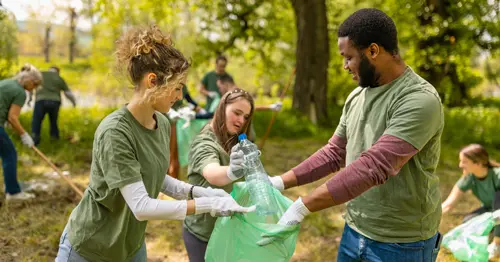 Three people wearing green shirts and gloves work together to collect plastic bottles in a green garbage bag during a park cleanup, with others picking up litter in the background surrounded by trees.